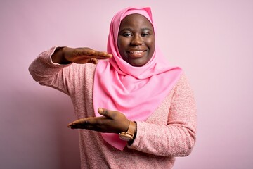 Young african american plus size woman wearing muslim hijab over isolated pink background gesturing with hands showing big and large size sign, measure symbol. Smiling looking at the camera. Measure