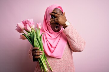 Young african american plus size woman wearing muslim hijab holding bouquet of pink tulips peeking in shock covering face and eyes with hand, looking through fingers with embarrassed expression.