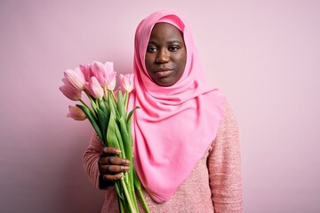 Young african american plus size woman wearing muslim hijab holding bouquet of pink tulips Relaxed with serious expression on face. Simple and natural looking at the camera.