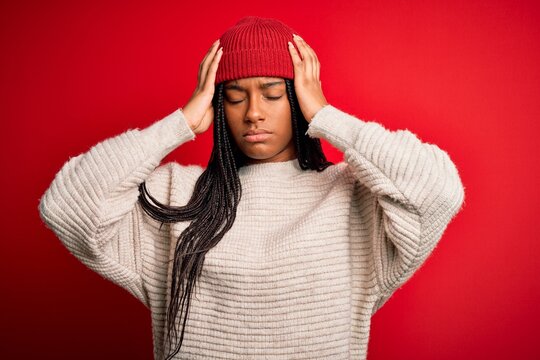 Young african american woman wearing winter sweater and wool hat over red isolated background suffering from headache desperate and stressed because pain and migraine. Hands on head.