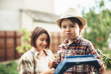 Charming caucasian boy with a hat on head eating cherries from the tree posing with his mother on background smiling