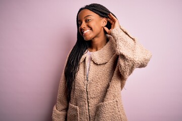 Young african american woman wearing fashion winter coat over pink isolated background smiling with hand over ear listening an hearing to rumor or gossip. Deafness concept.