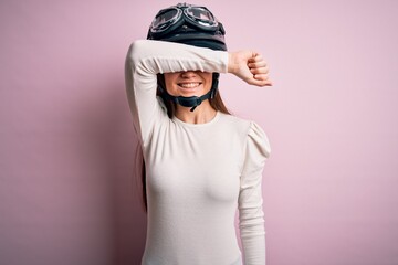 Young beautiful motorcyclist woman with blue eyes wearing moto helmet over pink background Smiling cheerful playing peek a boo with hands showing face. Surprised and exited