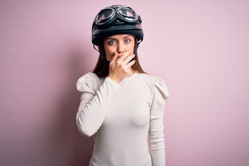 Young beautiful motorcyclist woman with blue eyes wearing moto helmet over pink background shocked covering mouth with hands for mistake. Secret concept.
