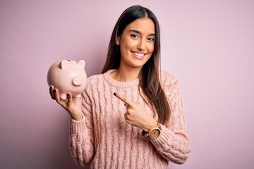 Young beautiful brunette woman holding piggy bank saving money for retirement very happy pointing with hand and finger