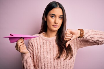 Young beautiful brunette woman holding paper airplane over isolated pink background with angry...