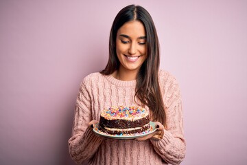 Young beautiful brunette woman holding birthday delicious cake over pink background with a happy face standing and smiling with a confident smile showing teeth