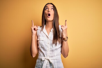 Young beautiful brunette woman on vacation wearing casual dress over yellow background amazed and surprised looking up and pointing with fingers and raised arms.