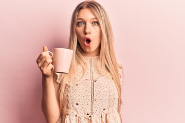 Young beautiful blonde woman holding coffee scared and amazed with open mouth for surprise, disbelief face