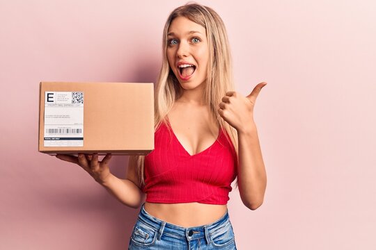 Young beautiful blonde woman holding delivery package pointing thumb up to the side smiling happy with open mouth