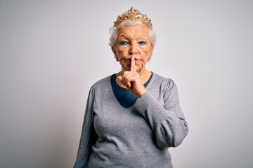 Senior beautiful grey-haired woman wearing golden queen crown over white background asking to be quiet with finger on lips. Silence and secret concept.