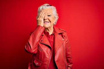 Senior beautiful grey-haired woman wearing casual red jacket and sunglasses covering one eye with hand, confident smile on face and surprise emotion.