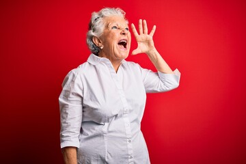 Senior beautiful woman wearing elegant shirt standing over isolated red background shouting and screaming loud to side with hand on mouth. Communication concept.
