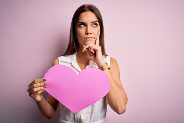 Young beautiful brunette woman holding big pink heart paper celebrating valentine day serious face thinking about question, very confused idea