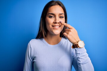 Young beautiful brunette woman wearing casual sweater standing over blue background Pointing with hand finger to face and nose, smiling cheerful. Beauty concept