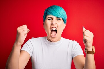 Young beautiful woman with blue fashion hair wearing casual t-shirt over red background angry and mad raising fists frustrated and furious while shouting with anger. Rage and aggressive concept.