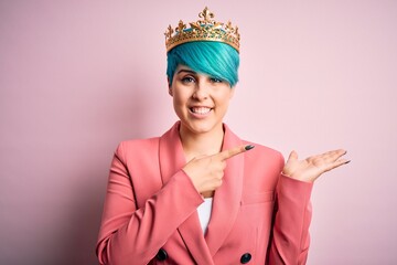 Young business woman with blue fashion hair wearing queen crown over pink isolated background amazed and smiling to the camera while presenting with hand and pointing with finger.