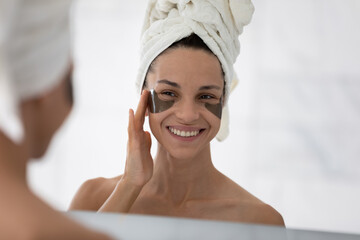 Mirror reflection smiling woman wearing white bath towel on head applying hydrogel eye care patches, standing in bathroom, moisturizing skin under eyes, enjoying skincare procedures