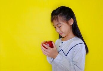 A cute young Asian girl holding up an apple, getting ready to eat it for breakfast. Bright yellow background.