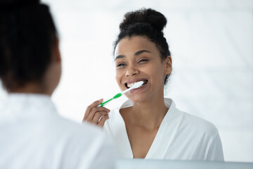Mirror reflection head shot close up smiling African American young woman brushing teeth, standing in bathroom, attractive girl with toothy smile cleaning teeth, morning routine, oral hygiene concept