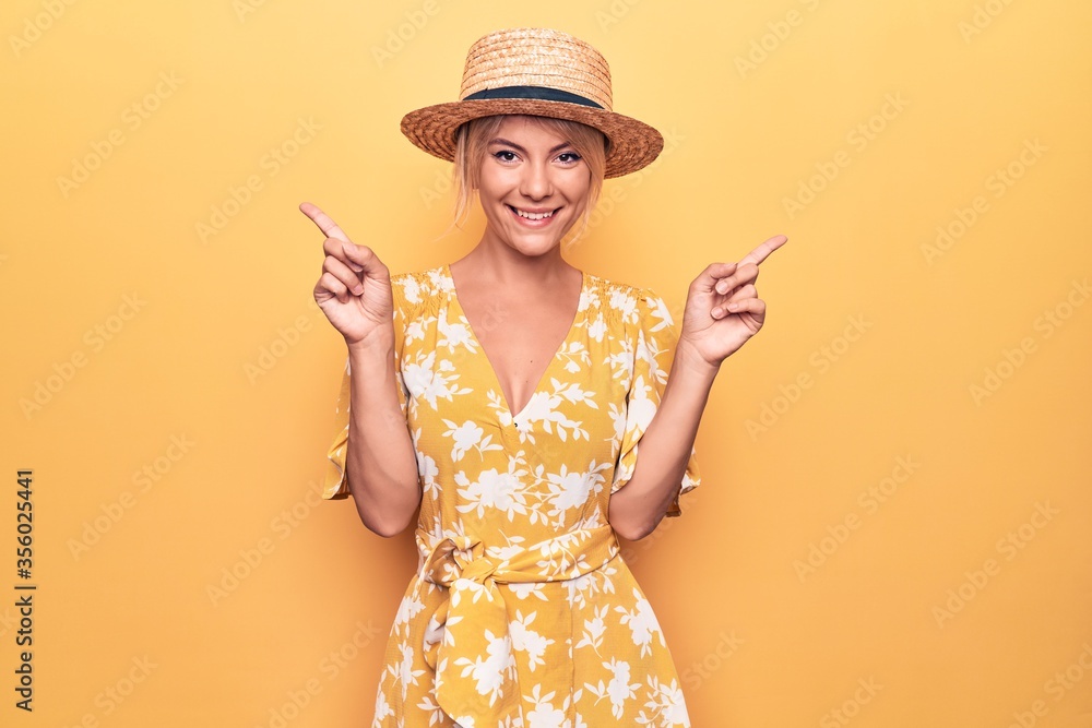 Wall mural beautiful blonde woman on vacation wearing summer hat and dress over yellow background smiling confi