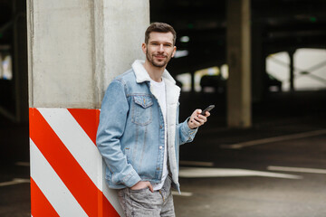 man in a fashion jacket holds a phone posing near a pillar in a city parking lot