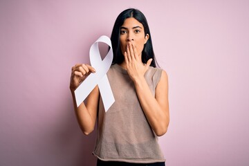 Young beautiful hispanic woman holding white ribbon as stop violence and lung cancer symbol cover mouth with hand shocked with shame for mistake, expression of fear, scared in silence, secret concept