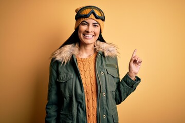 Young beautiful hispanic woman wearing ski glasses and coat for winter weather with a big smile on face, pointing with hand finger to the side looking at the camera.