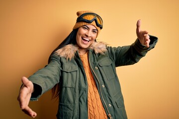 Young beautiful hispanic woman wearing ski glasses and coat for winter weather looking at the camera smiling with open arms for hug. Cheerful expression embracing happiness.