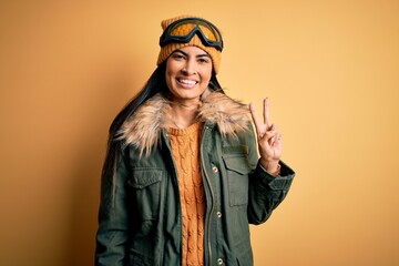 Young beautiful hispanic woman wearing ski glasses and coat for winter weather smiling looking to the camera showing fingers doing victory sign. Number two.