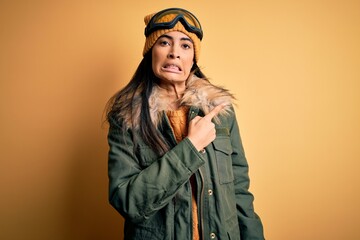 Young beautiful hispanic woman wearing ski glasses and coat for winter weather Pointing aside...