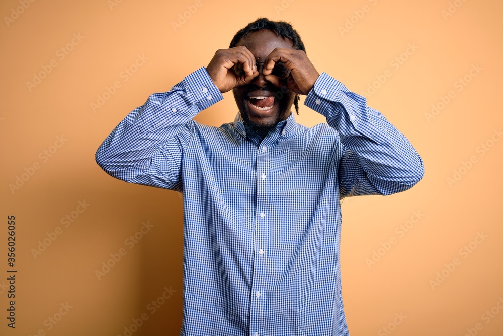 Poster Young handsome african american man wearing shirt and glasses over yellow background doing ok gesture like binoculars sticking tongue out, eyes looking through fingers. Crazy expression.