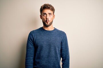 Young handsome man with beard wearing casual sweater standing over white background making fish face with lips, crazy and comical gesture. Funny expression.
