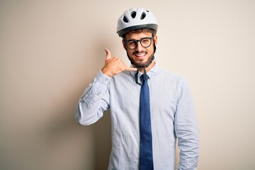 Young businessman wearing glasses and bike helmet standing over isolated white bakground smiling doing phone gesture with hand and fingers like talking on the telephone. Communicating concepts.