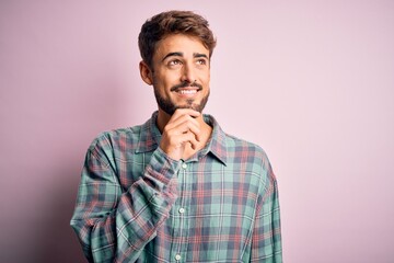 Young handsome man with beard wearing casual shirt standing over pink background with hand on chin thinking about question, pensive expression. Smiling with thoughtful face. Doubt concept.