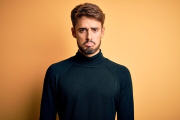 Young handsome man with beard wearing turtleneck sweater standing over yellow background depressed and worry for distress, crying angry and afraid. Sad expression.