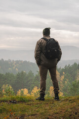Fototapeta na wymiar A man traveler with a backpack stands on the edge of the mountain and looks at the autumn forest in rainy weather.