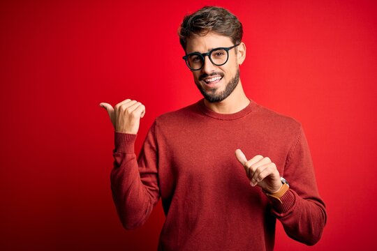 Young handsome man with beard wearing glasses and sweater standing over red background Pointing to the back behind with hand and thumbs up, smiling confident