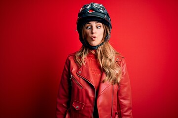 Young beautiful brunette motrocyclist woman wearing moto helmet over red background making fish face with lips, crazy and comical gesture. Funny expression.
