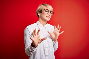 Young blonde business woman with short hair wearing glasses over red background disgusted expression, displeased and fearful doing disgust face because aversion reaction. With hands raised