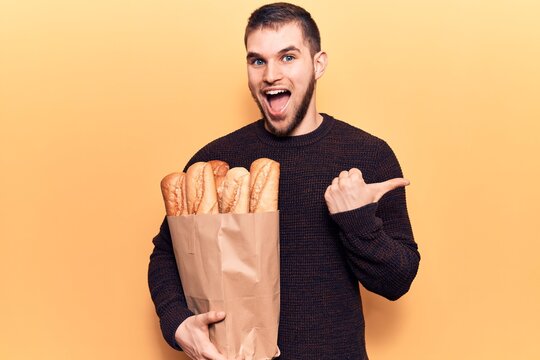 Young handsome man holding paper bag with bread pointing thumb up to the side smiling happy with open mouth