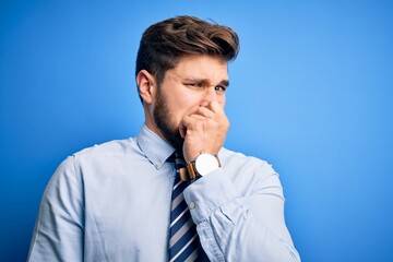 Young blond businessman with beard and blue eyes wearing elegant shirt and tie standing smelling something stinky and disgusting, intolerable smell, holding breath with fingers on nose. Bad smell