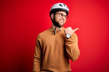 Young blond cyclist man with beard and blue eyes wearing bike helmet and glasses smiling with happy face looking and pointing to the side with thumb up.