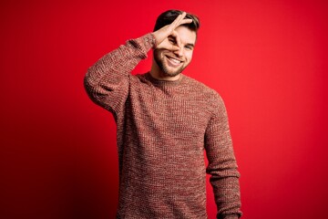 Young blond man with beard and blue eyes wearing casual sweater over red background doing ok gesture with hand smiling, eye looking through fingers with happy face.