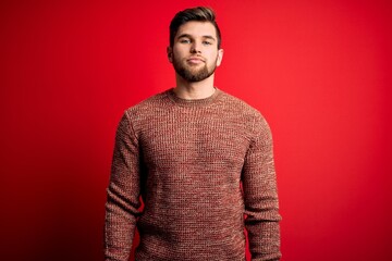 Young blond man with beard and blue eyes wearing casual sweater over red background with serious expression on face. Simple and natural looking at the camera.