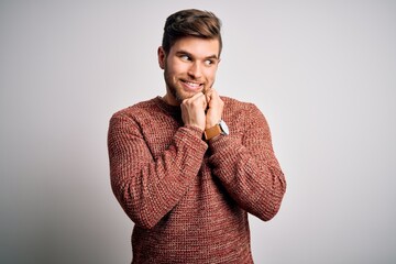 Young blond man with beard and blue eyes wearing casual sweater over white background laughing nervous and excited with hands on chin looking to the side