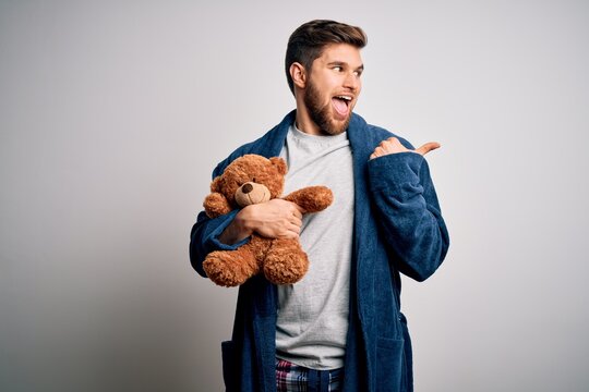 Young blond man with beard and blue eyes wearing pajama holding teddy bear pointing and showing with thumb up to the side with happy face smiling