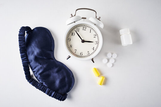 Insomnia, Depression And Sleeping Trouble Problem Concept. Alarm Clock, Sleeping Mask, Ear Plugs And Pills On A White Background, Top View