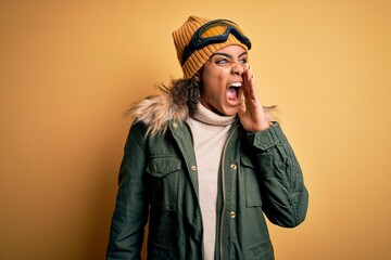 Young african american afro skier girl wearing snow sportswear and ski goggles shouting and screaming loud to side with hand on mouth. Communication concept.