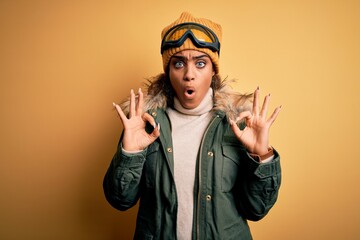 Young african american afro skier girl wearing snow sportswear and ski goggles looking surprised and shocked doing ok approval symbol with fingers. Crazy expression